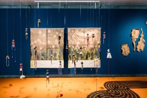 Ovadali vounona (Birthers of children) I (2023), painting, silk fabrics, linen fabric, archival image transfer, cotton embroidery threads, paint markers, acrylic paint, gold leafing marker, copper leafing marker, 270 x 217 cm; Ovadali vounona (Birthers of children) Il (2023), painting, silk fabrics, linen fabrics, archival image transfer, cotton embroidery threads, paint markers, acrylic paint, gold leafing marker, copper leafing marker, 270 x 217 cm; Ounona vedu (Children of the soil) (2023), installation, wood, antique glass beads, shell, shell beads, cowrie shells, copper beads, seeds, safety pins, human hair, nails, brass beads, suede, leather, springbok fur and skin, buttons, metal beads, ostrich egg shell beads, suede, cotton yarn, cotton fabric, linen fabric, rusted metal rings, sticks, red ochre, modelling clay, dimensions vary. Courtesy of the artist. Installation view of the exhibition, Haus der Kulturen der Welt (HKW), 2023.&nbsp;
